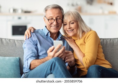 Happy Senior Couple Using Smartphone Browsing Internet Together Sitting On Couch At Home And Hugging. Cellphone Users Use Mobile Application. Older Spouses Texting On Phone. Communication Concept