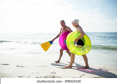 Happy senior couple walking on beach with inflatable rings and flippers on a sunny day