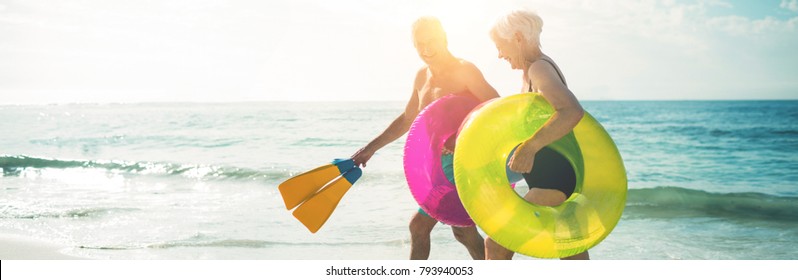 Happy senior couple walking on beach with inflatable rings and flippers during sunny day - Powered by Shutterstock