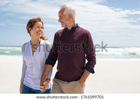 Happy senior couple walking on the beach in a sunny day. Smiling mature couple looking at each other on beach during sunset with copy space. Retired man in love with his wife relaxing during vacation.