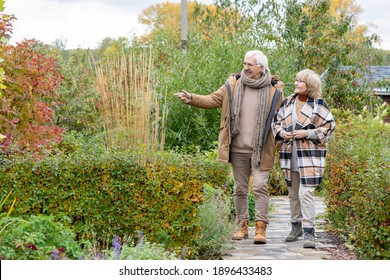 Happy Senior Couple In Warm Casualwear Moving Down Narrow Road Between Bushes And Trees While Discussing Plants In Their Garden