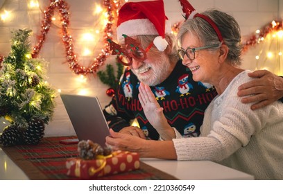 Happy Senior Couple Wearing Santa Hat At Christmas Time In Video Call By Laptop From Home Getting In Touch With Family Or Friends