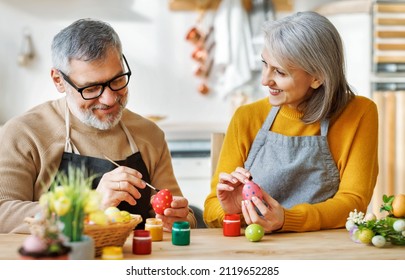 Happy senior couple wife and husband sit at table in kitchen, dye and decorate boiled eggs with food paints, mature family prepare together for Easter holiday and joyfully talking, selective focus - Powered by Shutterstock