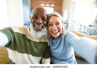 Happy Senior Diverse Couple In Living Room Sitting On Sofa, Making Video Call. Retirement Lifestyle, At Home With Technology.