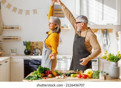 Happy senior elderly couple husband and wife joyfully smiling, tenderly embracing or dancing while cooking together in kitchen, senior family having fun while preparing vegetarian dishes - Powered by Shutterstock