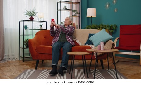 Happy senior elderly grandfather user holding smartphone watching mobile video calling online looking at screen making selfie, relaxing on couch at home. Old man learn using modern technology device - Powered by Shutterstock