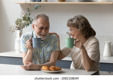 Happy senior family couple eating bakery food for breakfast in home kitchen, enjoying morning coffee, drinking mugs of hot tea, talking, smiling, laughing. Elderly age, marriage, love concept - Powered by Shutterstock