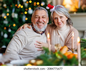 Happy senior family couple, grandparents, sitting at dinning table near decorated christmas tree at home and smiling at camera, positive mature man and woman embracing during New Year celebration - Powered by Shutterstock