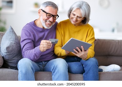 Happy Senior Family Couple Paying Bills Online With Credit Card, Retired Husband And Wife Using Digital Tablet For Shopping And Buying Good In Internet While Sittinng On Sofa In Living Room At Home