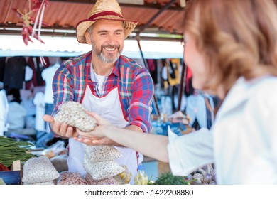 Happy Senior Farmer Standing Behind Market Stall, Selling Organic Beans And Vegetables In A Marketplace. Serving Mature Female Customer. Healthy Organic Local Food Production