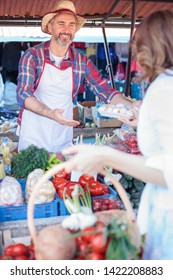 Happy Senior Farmer Standing Behind Market Stall, Selling Organic Vegetables In A Farmer's Marketplace. Serving Mature Female Customer. Healthy Organic Local Food Production