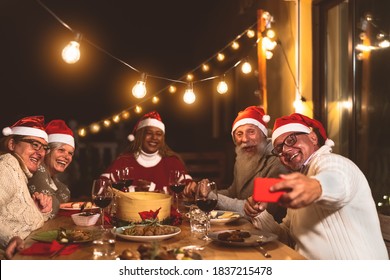 Happy senior friends dining and celebrating Christmas holidays while taking selfie with mobile smartphone on house patio party