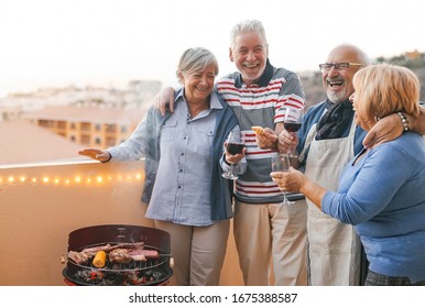 Happy senior friends having fun cheering with red wine at barbecue on terrace  - Mature people dining and laughing together at bbq party - Food drink and elderly friendship lifestyle concept