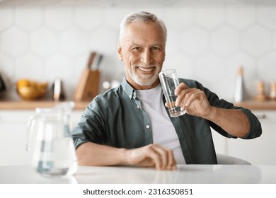 Happy Senior Gentleman Holding Glass With Water While Sitting At Table In Kitchen, Smiling Elderly Man Enjoying Refreshing Drink And Looking At Camera, Thirsty Male Taking Healthy Liquid - Powered by Shutterstock