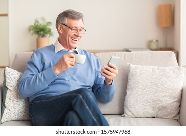 Happy Senior Gentleman Using Smartphone Texting And Having Coffee Sitting On Sofa At Home. Selective Focus