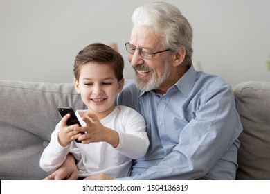 Happy senior grandfather and cute small grandson using smart phone apps at home, old grandpa embracing little grandchild holding looking at cellphone screen playing mobile game relaxing sit on sofa