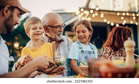 Happy Senior Grandfather Talking and Having Fun with His Grandchildren, Holding Them on Lap at a Outdoors Dinner with Food and Drinks. Adults at a Garden Party Together with Kids. - Powered by Shutterstock