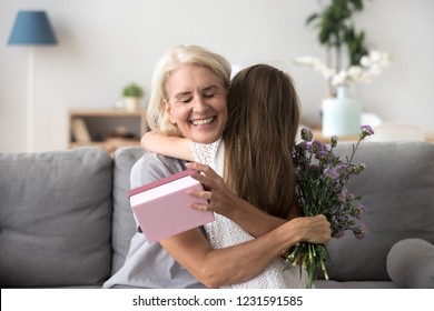 Happy Senior Grandma Hugging Granddaughter Thanking For Present Holding Flower Bouquet, Smiling Excited Old Grandmother Embracing Little Grandchild Girl Congratulating Granny Giving Birthday Gift Box