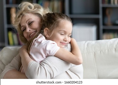 Happy Senior Grandmother Sit On Couch In Living Room Hugging Cute Little Preschooler Granddaughter, Smiling Mature 70s Granny Embrace Cuddle With Small Grandchild, Show Love And Care, Bonding Concept