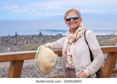 Happy senior gray haired woman with backpack walking along the sea beach with hat in hand enjoying vacation, free time or retirement - Shutterstock ID 2160165855