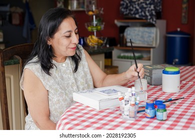 Happy senior Hispanic woman doing a craft at home - woman smiling while painting with paintbrush on wooden box