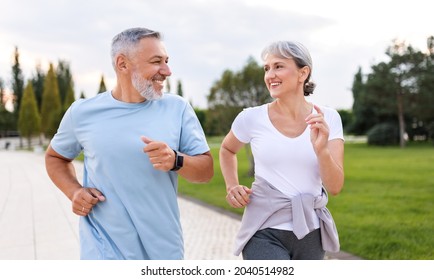 Happy Senior Husband And Wife In Sportive Outfits Running Outdoors In City Park, Lovely Retirees Couple Jogging In Sunny Morning Looking At Each Other With Warmth And Smile. Healthy Lifestyle Concept