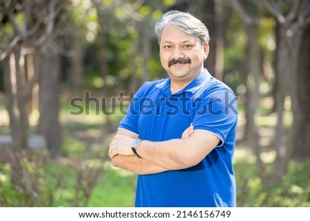 Happy senior indian asian man with arms crossed standing at park outdoor, Old mustache and grey haired male smiling wearing blue t-shirt. Copy space.