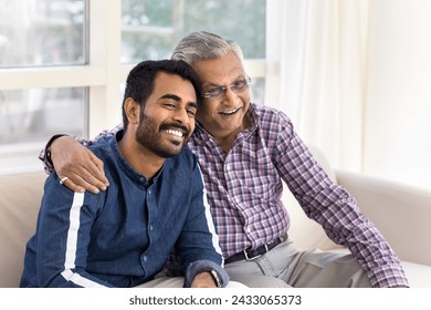 Happy senior Indian dad hugging positive adult son with love, support, pride, looking away with perfect toothy smile, laughing enjoying family meeting, home leisure, sitting on couch - Powered by Shutterstock