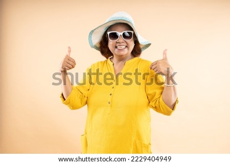 Happy senior indian woman wearing hat and sunglasses do thumbs with both hands standing isolated over beige studio background, Retirement life, summer holidays and travel.
