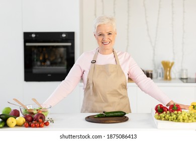 Happy Senior Lady Cooking In Kitchen Making Salad For Dinner At Home, Smiling To Camera. Aged Woman Preparing Healthy Meal Posing Indoors. Nutrition And Recipes For Older Female