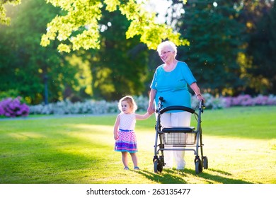 Happy senior lady with a walker or wheel chair and a little toddler girl, grandmother and granddaughter, enjoying a walk in the park. Child supporting disabled grandparent.