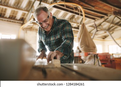 Happy Senior Male Carpenter Cutting Wood Planks On Table Saw Machine. Smiling Mature Man Working In Carpentry Workshop.