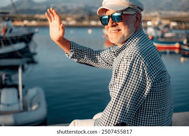 Happy senior man in casual shirt and hat sitting at the sea port greeting friends. Relaxed elderly bearded male wearing sunglasses enjoying freedom, travel or retirement - Powered by Shutterstock