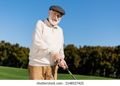 Happy Senior Man In Flat Cap Holding Golf Club While Playing Outdoors