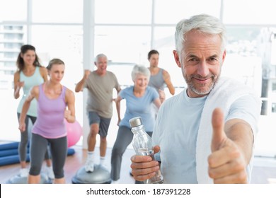 Happy senior man gesturing thumbs up with people exercising in the background at fitness studio