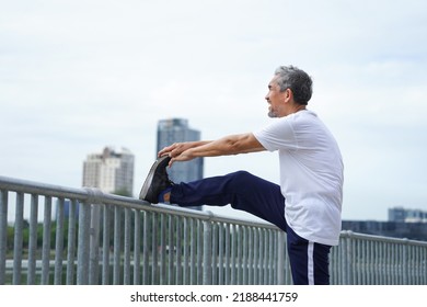 Happy Senior Man With Grey Hair Stretching Legs In The City Park, An Old Man Warming Up Before Workout To Prevent Injury. Concept For Elderly People Lifestyle, Health Care, Wellbeing