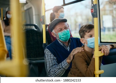 Happy Senior Man And His Grandson Traveling By Public Bus And Wearing Protective Face Masks Due To Coronavirus Pandemic.