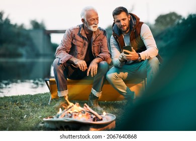 Happy senior man and his son using mobile phone while camping and relaxing by campfire. Copy space. - Powered by Shutterstock