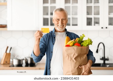 Happy Senior Man Holding Credit Card And Paper Bag With Groceries, Cheerful Elderly Gentleman Enjoying Easy Payments, Recommending Bank Services, Standing In Kitchen And Home And Smiling At Camera - Powered by Shutterstock