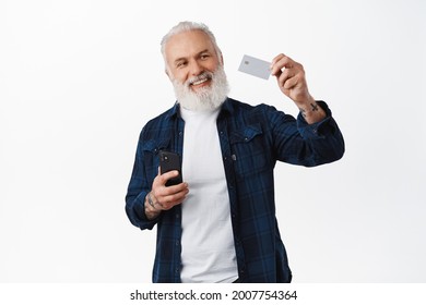 Happy senior man holding smartphone, raising hand and showing credit card with satisfied smile, nod in approval, recommend online payment, mobile banking app, standing over white background - Shutterstock ID 2007754364
