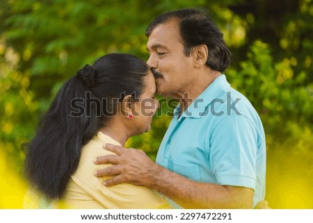 Happy senior man or husband kissing his wife's forehead by hugging at park - concept of emotional bonding, physical intimacy and romantic moment.