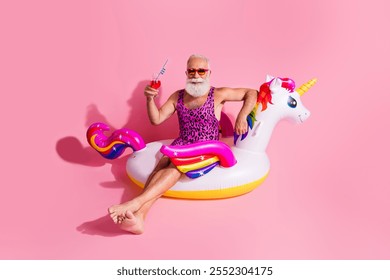 Happy senior man in leopard print swimwear enjoying a carefree summer day on an inflatable unicorn against a pink background - Powered by Shutterstock