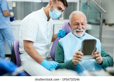 Happy Senior Man Looking His Teeth In A Mirror And Feeling Satisfied After Dental Appointment With His Dentist At Clinic. 