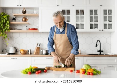 Happy senior man preparing healthy food in kitchen interior, smiling elderly gentleman cooking vegetarian lunch or dinner at home, mature male wearing apron chopping vegetables, copy space - Powered by Shutterstock