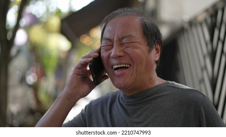 Happy Senior Man Receiving Great News On Phone Standing In City Street. Authentic Real Life Laugh And Smile Of An Older Asian Person Speaking With Cellphone Laughing