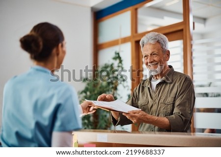 Happy senior man receiving his medical documents from nurse at reception desk at doctor's office.