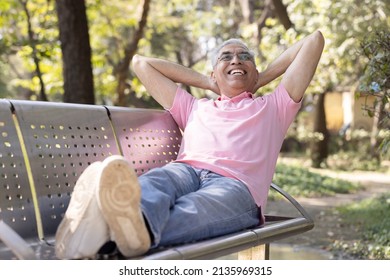 Happy Senior Man Relaxing With Hands Behind Head And Feet Up At Park Bench