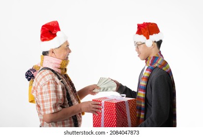 Happy Senior Man With Scarf And Santa Hat Give Money Or Dollar Bills To His Son, While His Son Give Giftbox For His Father For Celebrate Christmas Eve And Happy New Year On White Background.