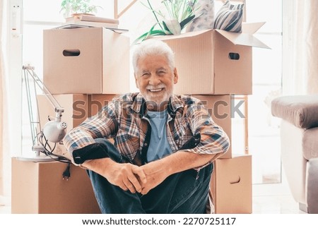 Happy senior man sitting on floor relaxing in new home living room with cardboard boxes packed with office stuff on moving day.