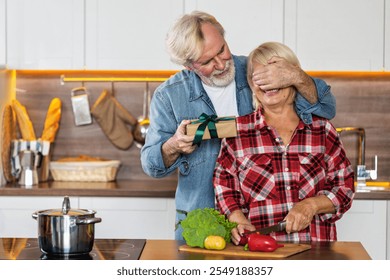 Happy senior man surprises his wife with a gift while she prepares vegetables in the kitchen at home. A moment of love and joy. Happy retirement concept - Powered by Shutterstock